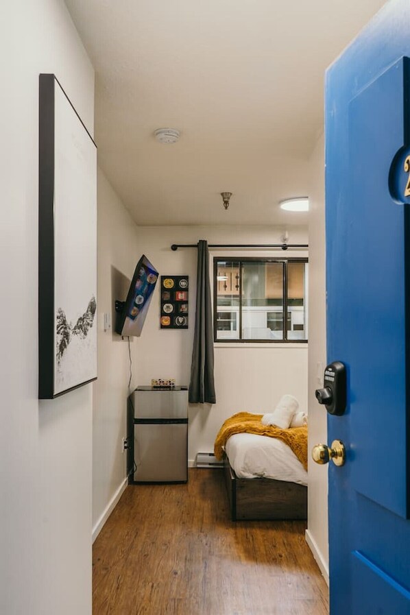 A compact bedroom with a bed, a small bedside table, a wall-mounted shelf, a door with a number plate, and a window with a view of the outside.