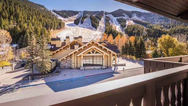 Eine Lodge mit Holzfassade, überdachter Parkmöglichkeit und Blick auf eine Skipiste im Hintergrund.