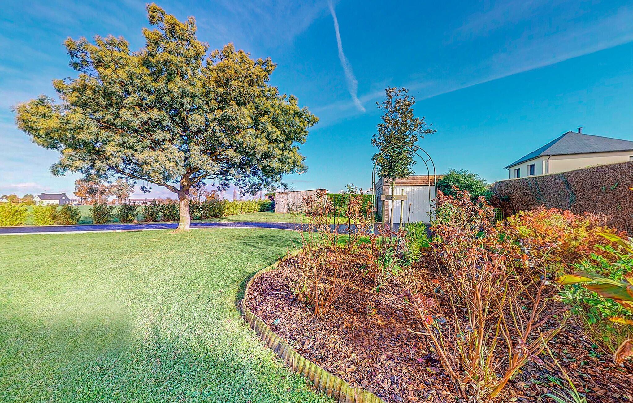A well-maintained garden with a large tree, a greenhouse, and a stone wall.