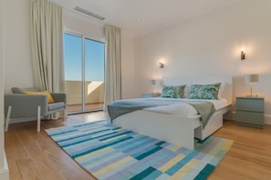 A modern bedroom with a large bed, a grey sofa, a colorful striped rug, and a view of the sky through the window.