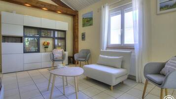 Una sala de estar con un sofá, dos sillones, una mesita redonda y una zona de cocina con armarios y una ventana.