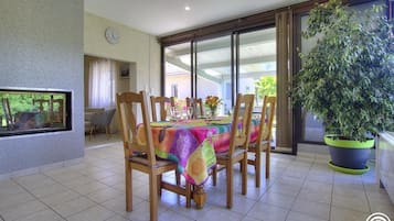 Un comedor con una mesa cubierta por un mantel colorido, rodeada de sillas de madera. A la derecha hay una planta grande en maceta y una ventana con vistas a follaje exterior.