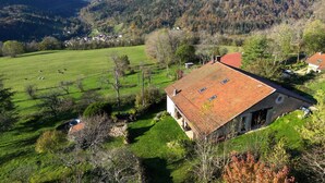 A rural house with a red roof, surrounded by green fields and trees, with a view of distant hills.