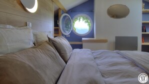 A bedroom with a bed, a round mirror, a window with a view, and a shelf with books.