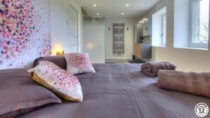 A modern bedroom with a large bed, a shower area, and a view of the kitchen.