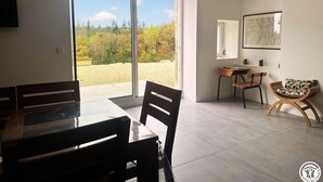 A modern dining area with a large table and chairs, a desk with a chair, and a view of a garden through a glass door.