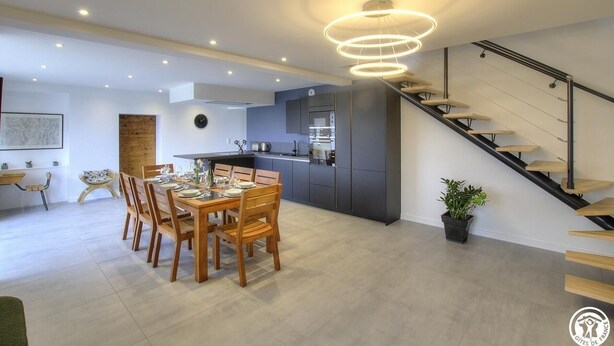 A modern kitchen with a central island, a dining area with wooden chairs, and a staircase with a minimalist design.