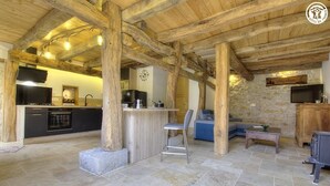 A rustic interior with exposed wooden beams, a stone wall, a kitchen area with modern appliances, and a seating area with a blue sofa and a small table.