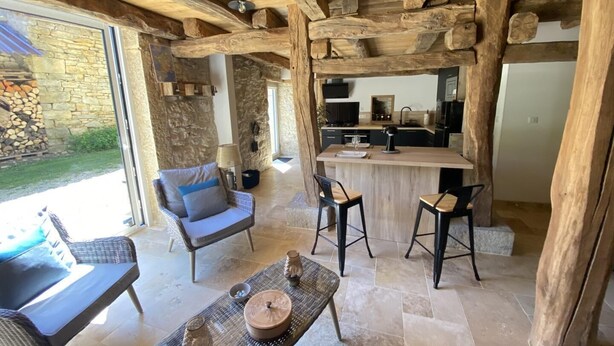 An open-plan living area with a stone wall, wooden ceiling beams, a kitchen, and a seating area with a table and chairs.