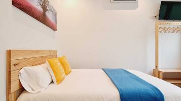 Une chambre d’hôtel avec une tête de lit en bois, un lit avec des oreillers blancs et jaunes, une couverture bleue, un climatiseur et un téléviseur posé sur un meuble en bois.