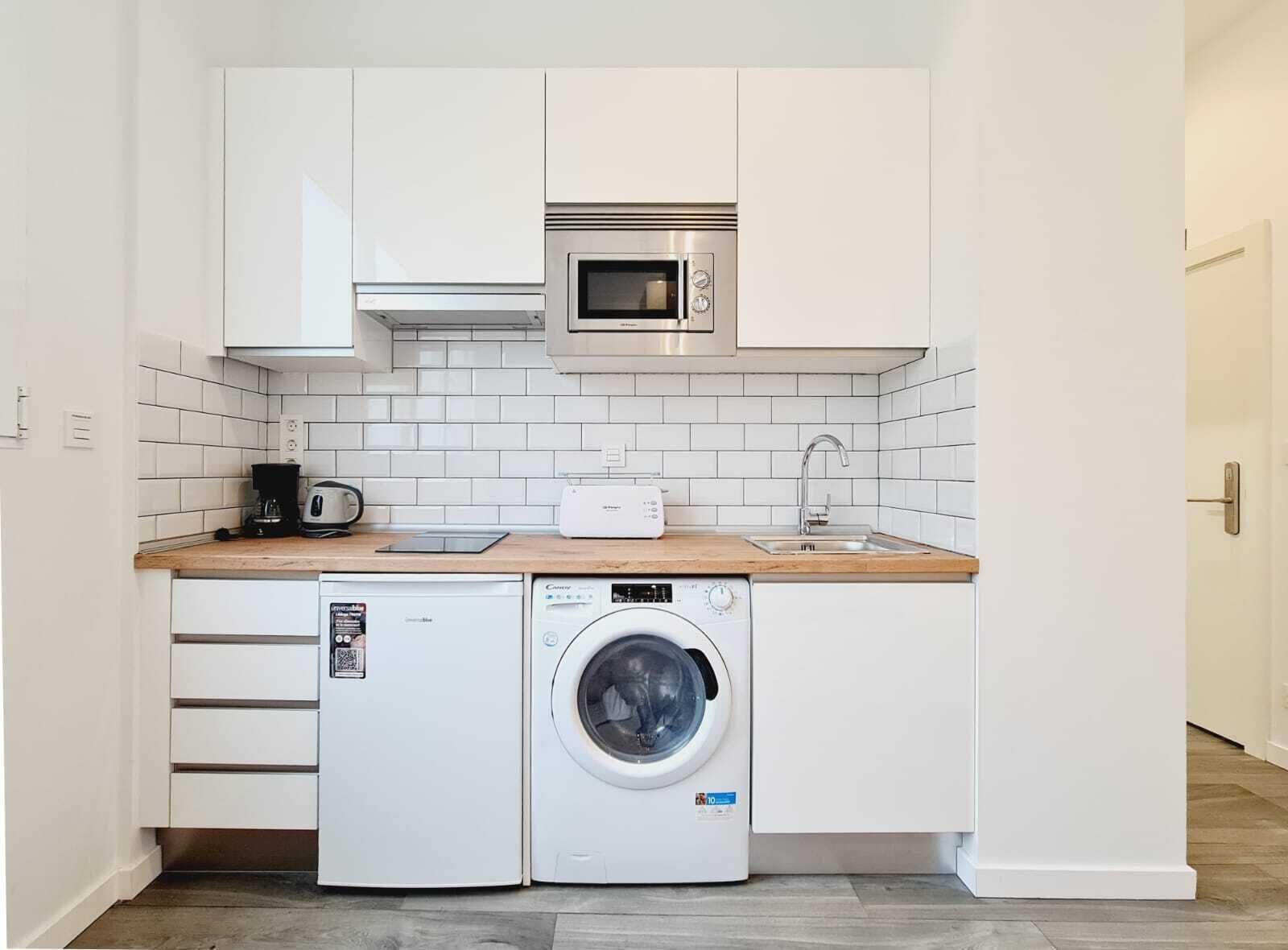 Una cucina compatta con mobili bianchi, un forno a microonde, una macchina da caffè, un tostapane e una lavatrice.