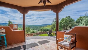 A covered patio with stone flooring, two wooden chairs with cushions, a birdcage, and a view of a green landscape with trees and mountains.