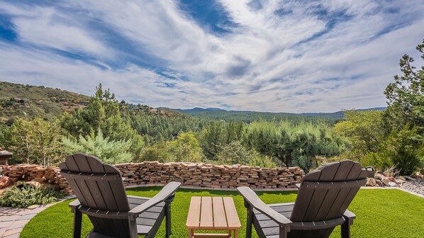 A patio with two Adirondack chairs and a small table, overlooking a forested valley with a stone wall and a grassy lawn.