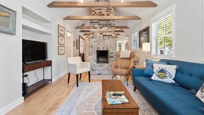 A living room with a brick fireplace, wooden beams, a blue sofa, and a television on a wooden stand. - Cozy Cottage across the street from Carl Sandburg National Park (Flat Rock)