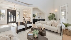 Un salón amplio con un sofá moderno, sillones oscuros y una mesa de centro. Se ve una zona de cocina al fondo.
