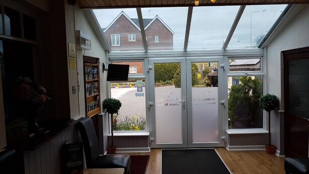 A reception area with a counter, seating, and a view of the street through glass doors. - Meadows Way Guest House (Uttoxeter)