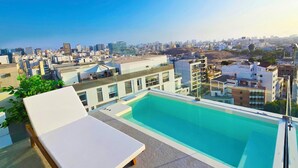 Una piscina en la azotea con vistas a la ciudad, sillas de descanso y edificios modernos al fondo.