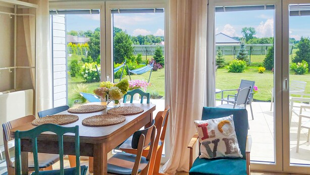 A dining area with a table set for four, surrounded by chairs, and large windows offering a view of a garden and a house. - Nice home in Sianow with sauna (Sianow)
