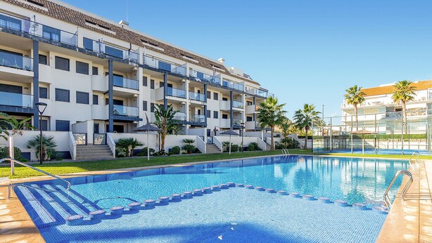 A swimming pool with a tiled deck, surrounded by a multi-story building with balconies, palm trees, and a clear sky. - 1 bedroom lovely apartment in Denia (Denia)