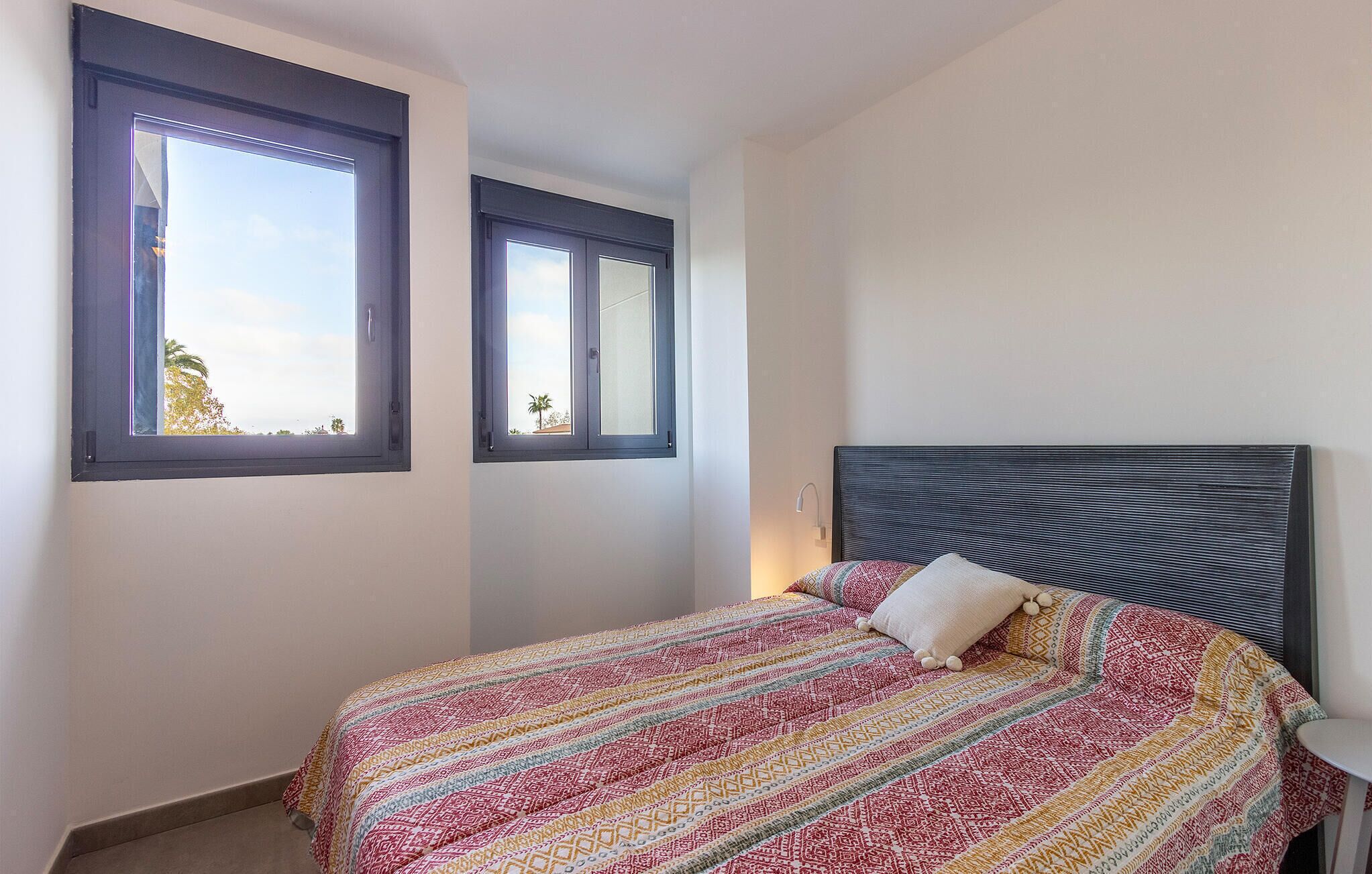 Ein Schlafzimmer mit einem Bett, zwei Fenstern, einem Nachttisch und einer Wandlampe.