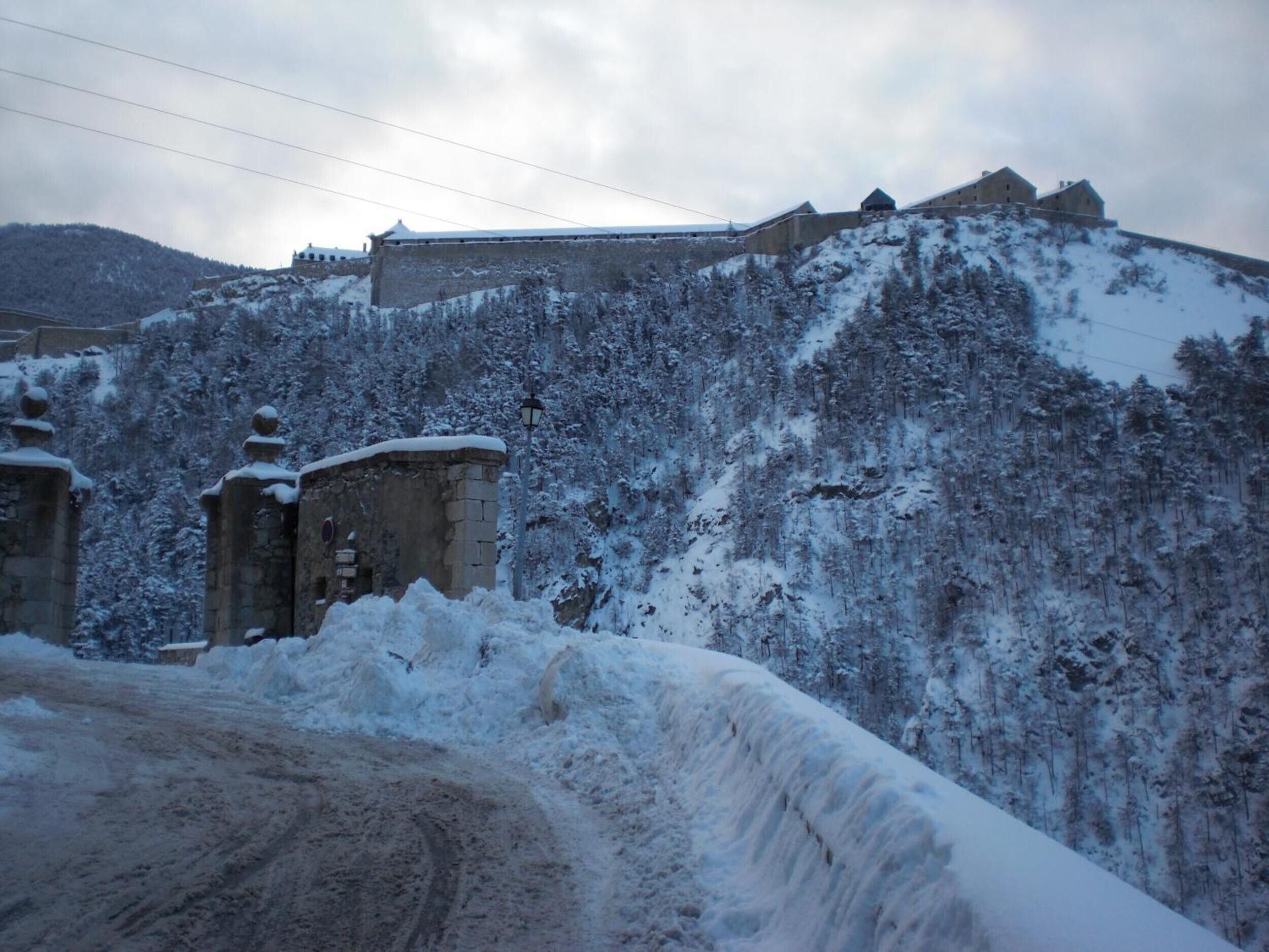 A snow-covered landscape with a stone wall and a building on a hill.