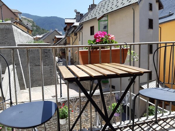 A balcony with a wooden table and two chairs, a potted plant with pink flowers, and a view of a street with buildings and a mountain in the background.