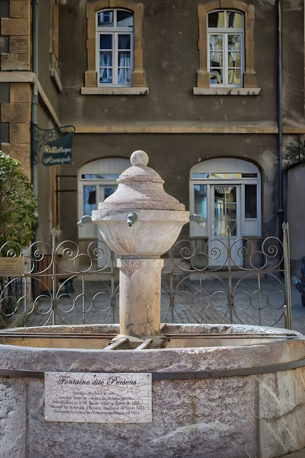 A stone fountain with a plaque, surrounded by a wrought iron fence, in front of a building with arched windows and a sign.