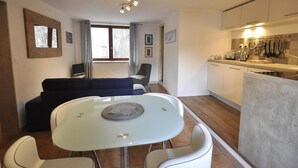 A modern kitchen with white cabinets, a dining area with a glass table, and a living area with a dark sofa. - Apartment 'Appartement Deux Chambres' with Mountain View, Private Terrace and Wi-Fi (Briançon)