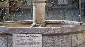 A stone fountain with a plaque in front of a building with arched windows and a sign above the entrance.