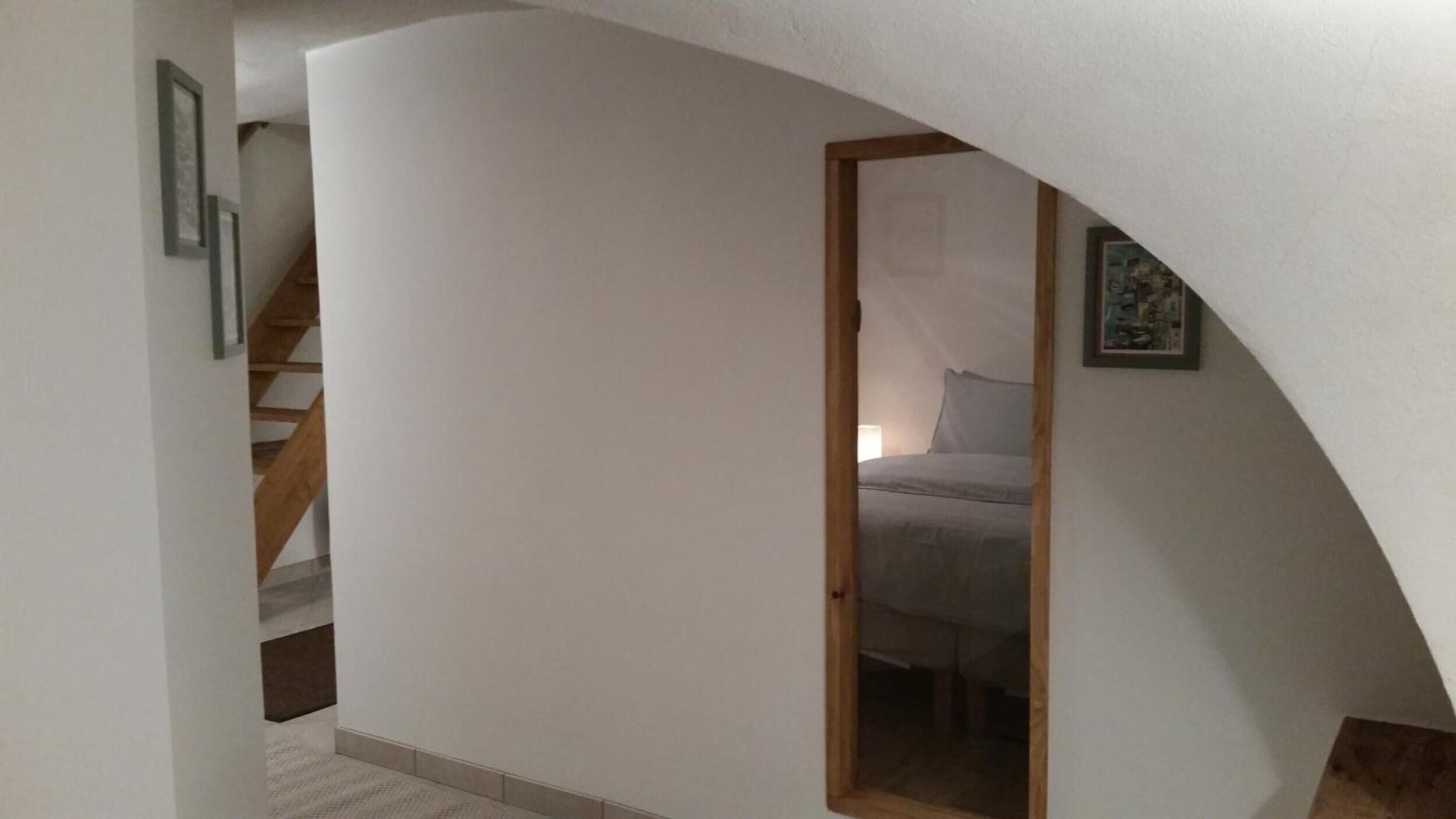 A narrow hallway with a wooden staircase on the left, a door leading to a bedroom with a bed and a nightstand, and a painting on the wall.