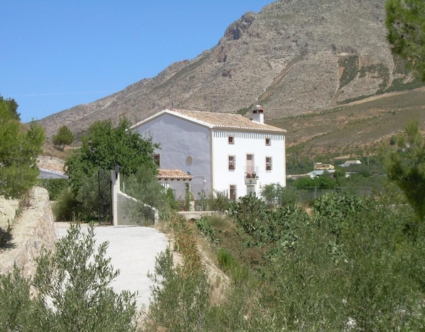 Un bâtiment blanc, doté d’un toit en tuiles, entouré de verdure et adossé à un paysage montagneux.