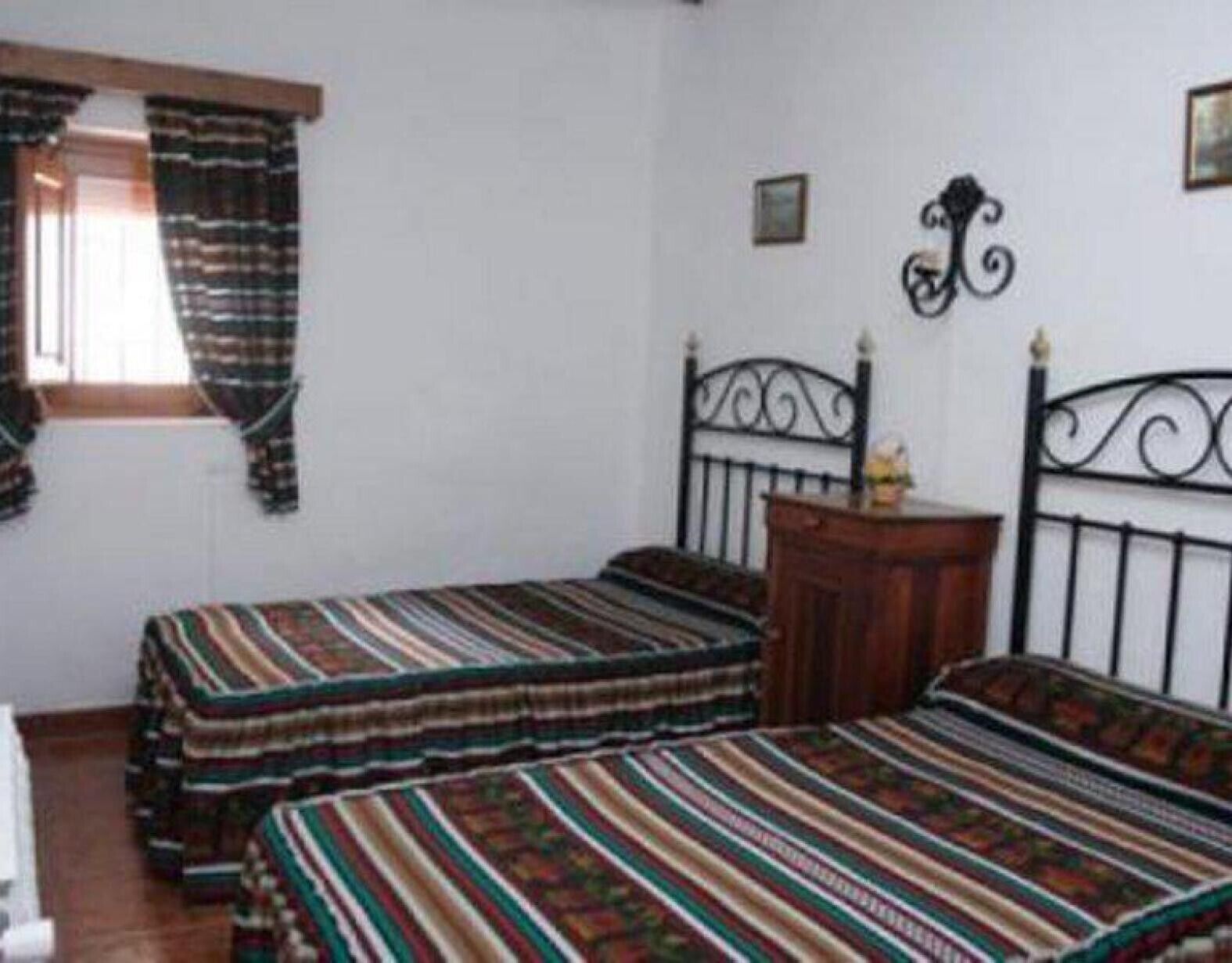 Une chambre avec deux lits simples, une table de chevet en bois, une tête de lit en métal noir et une fenêtre avec des rideaux.