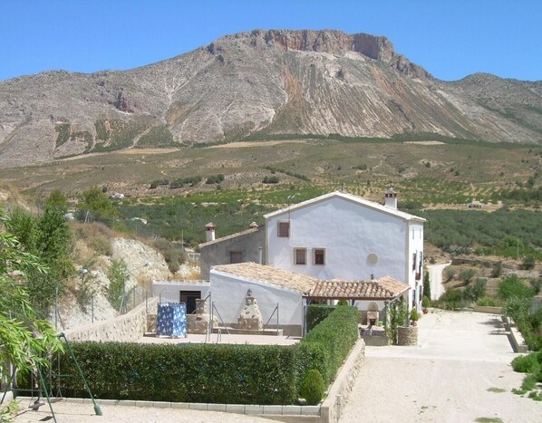 Un edificio blanco con techo de tejas, un pequeño jardín y una montaña al fondo.