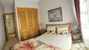 A bedroom with a bed, a wooden wardrobe, a nightstand, a lamp, and a painting on the wall.