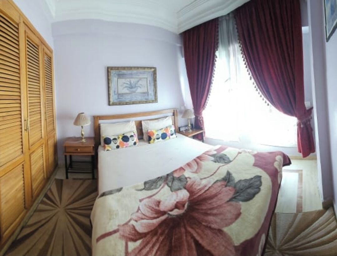 A bedroom with a large bed, wooden wardrobe, a nightstand, a lamp, a framed picture on the wall, and a window with curtains.
