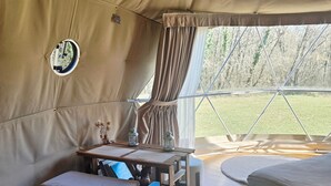A tent with a wooden table, two beds, a small table with books, and a view of trees outside.