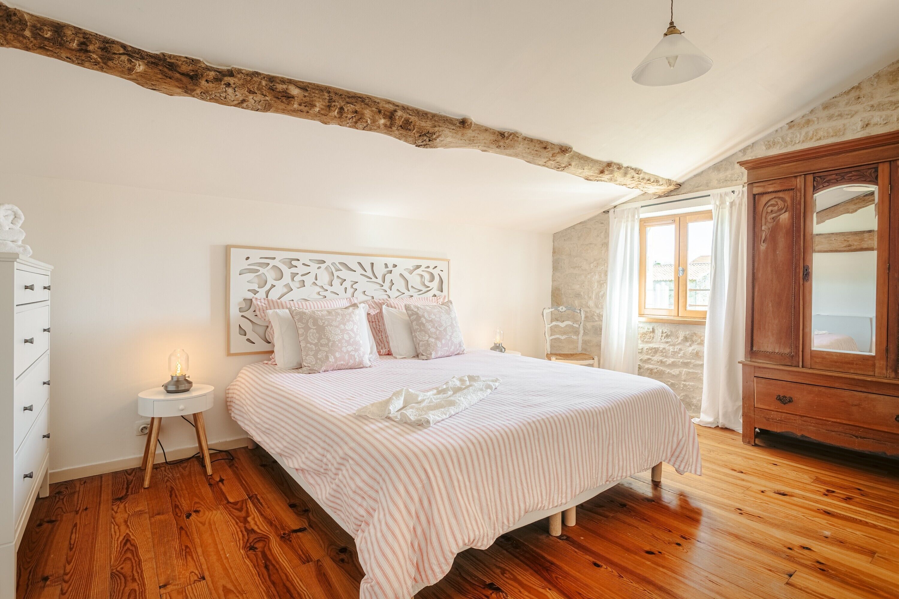 A bedroom with a wooden floor, a bed with a striped bedspread, a white nightstand with a lamp, a wooden wardrobe, and a stone wall.