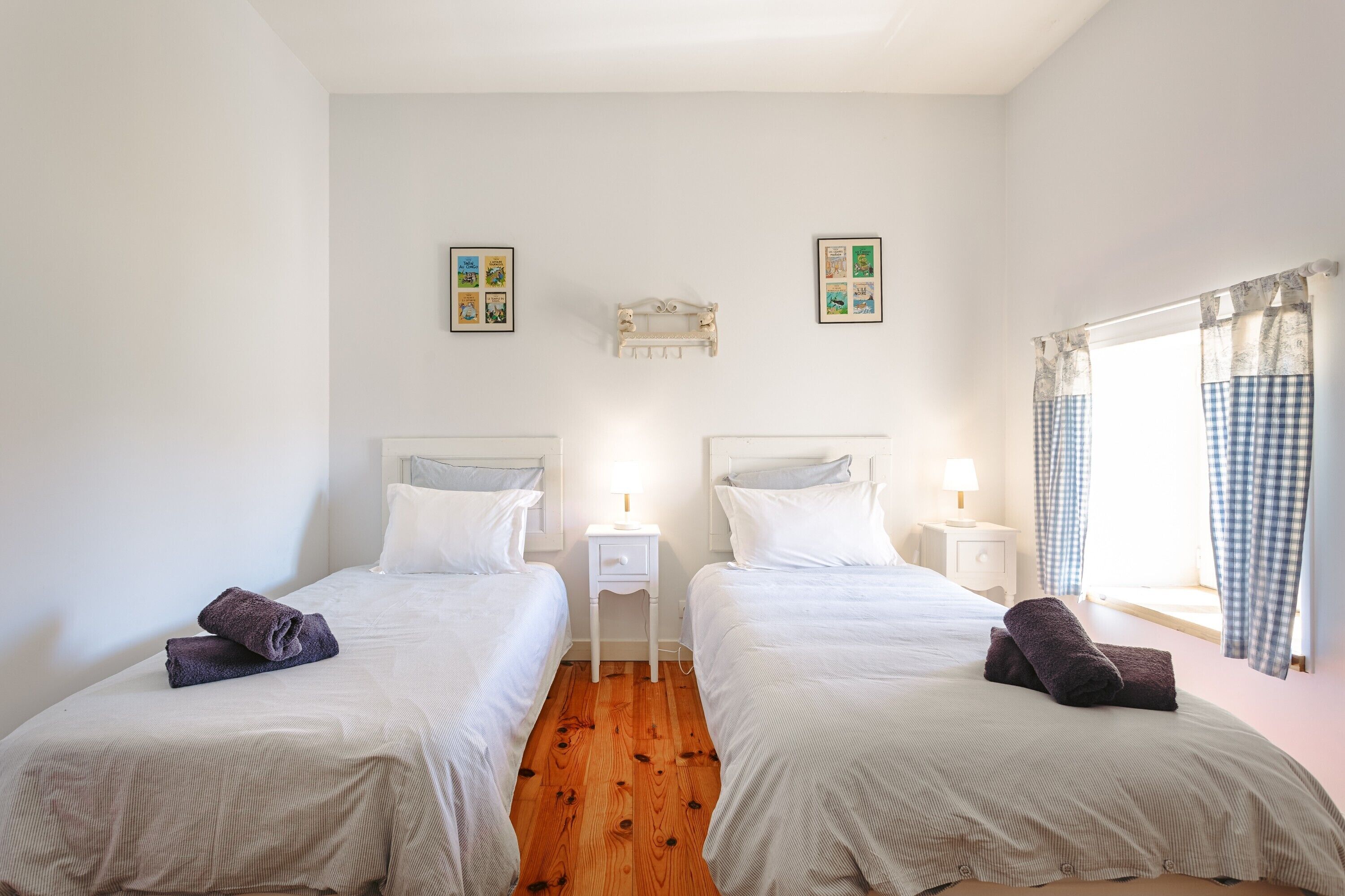 Una habitación con dos camas, una mesita de noche con lámpara, un estante en la pared y una ventana con cortinas.
