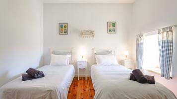 Una habitación con dos camas, una mesita de noche con lámpara, un estante en la pared y una ventana con cortinas.
