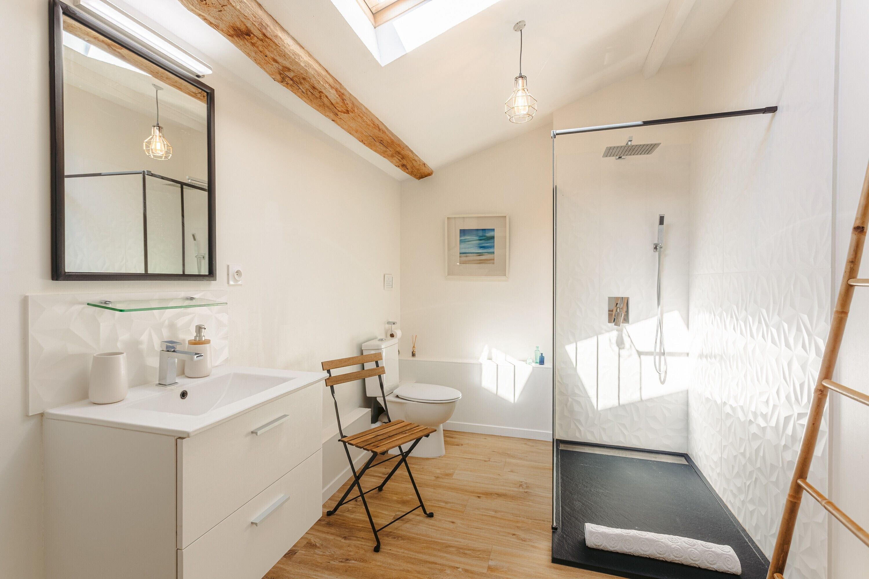 A modern bathroom with a skylight, a glass shower enclosure, a wooden chair, and a white sink.