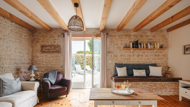A cozy living room with stone walls, wooden beams, a sofa, a coffee table, and a bookshelf. - Maison 'Ambre' with Shared Pool & Spa, Private Terrace, and Wi-Fi (Vergné)