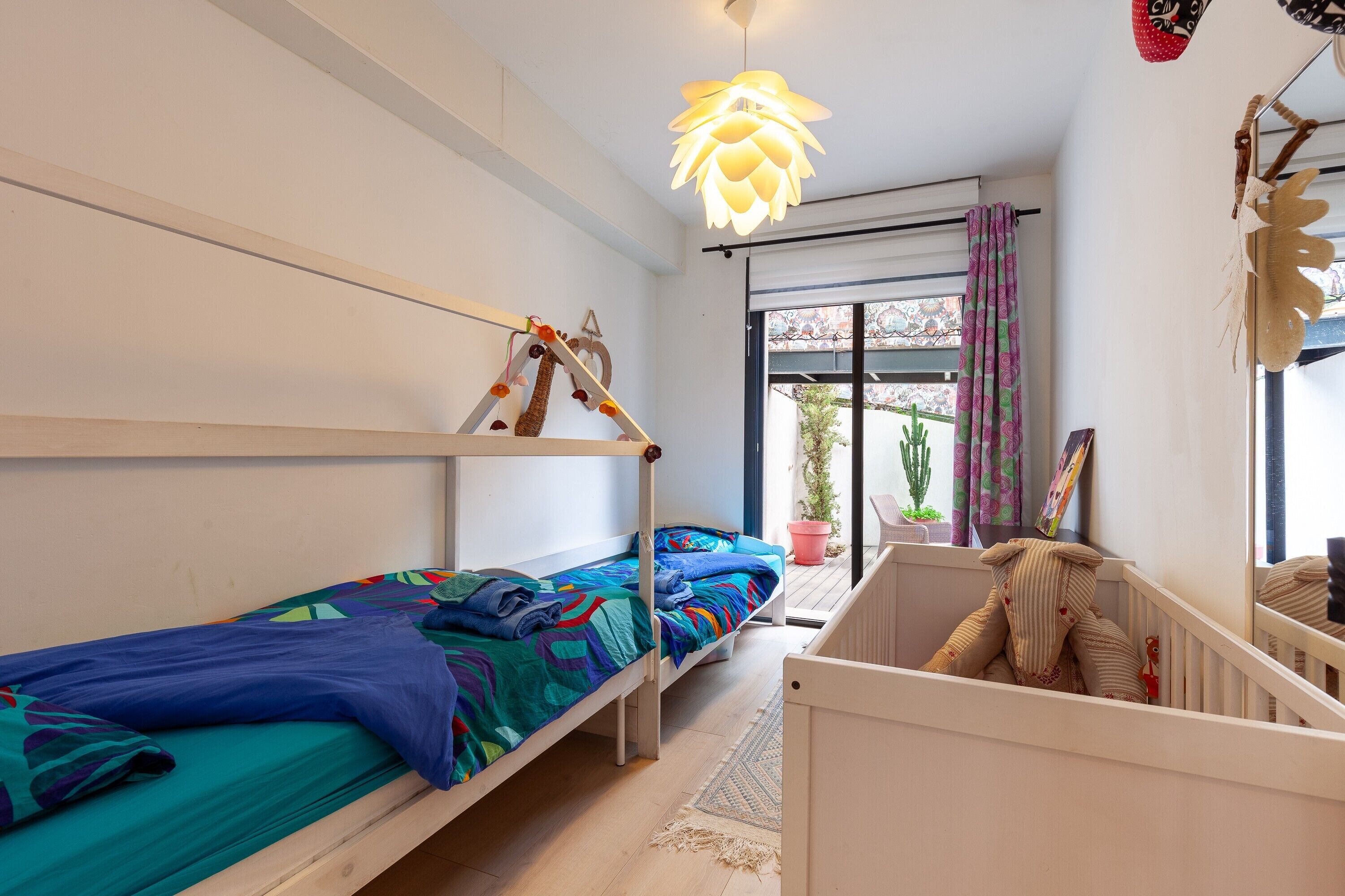 Een kamer met stapelbedden, een wiegje, een hanglamp, een raam met gordijnen en een plant in een pot.