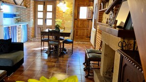 A cozy living room with a sofa, a dining table with chairs, a fireplace, and a kitchen area with a refrigerator and a window. - ‘La Tablá’ Rural Apartment with Mountain Views, Private Terrace, and Wi-Fi (Alles)