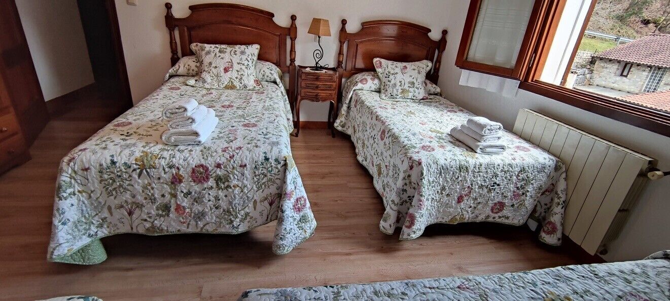 Een kamer met twee bedden, elk met bloemenbedrukking en handdoeken, een houten nachtkastje met een lamp, en een raam met uitzicht op gebouwen.