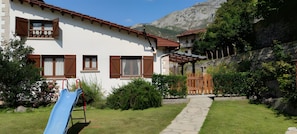 Une maison avec un chemin de pierre menant à un portail en bois, entourée de verdure et avec une chaîne de montagnes en arrière-plan.