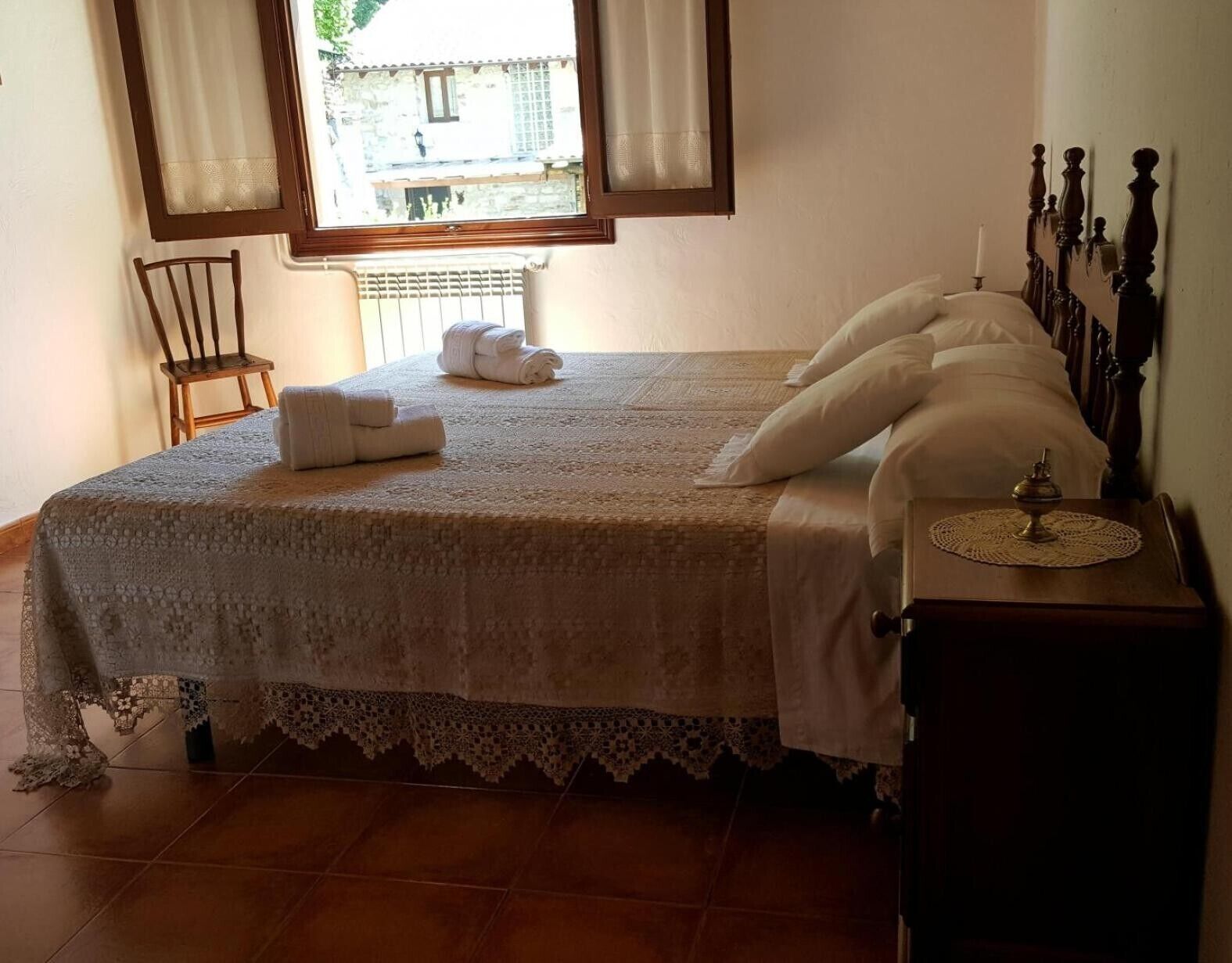 A neatly made bed with a lace bedspread, a wooden chair, a bedside table with a lamp, and a window with curtains.