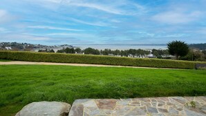 A grassy area with a stone pathway, a hedge, and a distant view of a body of water and buildings. - Villa 'Ker Louise' with Sea View, Shared Terrace and Wi-Fi (Locquirec)