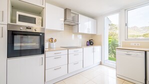 Een moderne keuken met witte kasten, een ingebouwde oven, magnetron en vaatwasser, en een groot raam met uitzicht op groen.