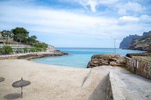 A beach with a stone wall, a clear blue sea, and a building with trees in the background. - 'Can Botana 10' with Sea View, Wi-Fi and Air Conditioning (Cala Sant Vicenç)