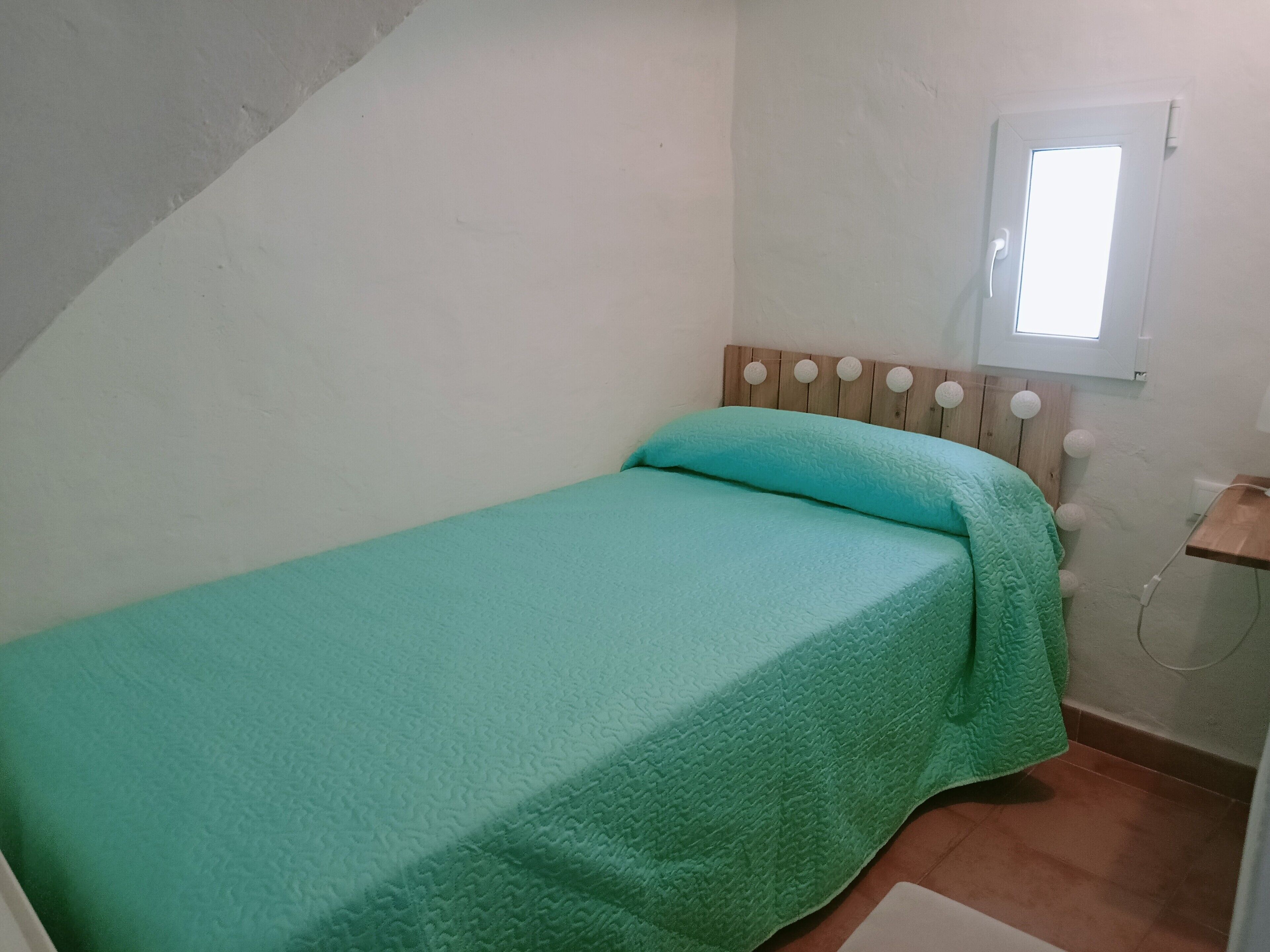 Une petite chambre avec un lit simple, un plafond en pente, une tête de lit en bois ornée de boutons ronds, une fenêtre donnant sur le ciel et un sol carrelé.